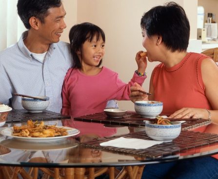 An Asian family, an adult male and female are seated around a table eating a meal with a young female standing in between the adults.