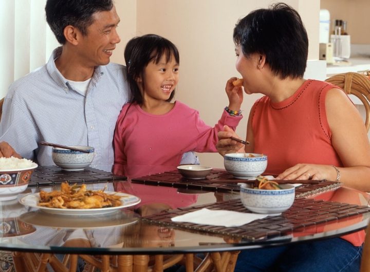 An Asian family, an adult male and female are seated around a table eating a meal with a young female standing in between the adults.
